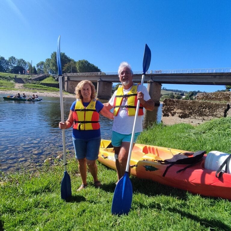 Pareja con chalecos salvavidas y remos junto a un kayak en la orilla de un río.