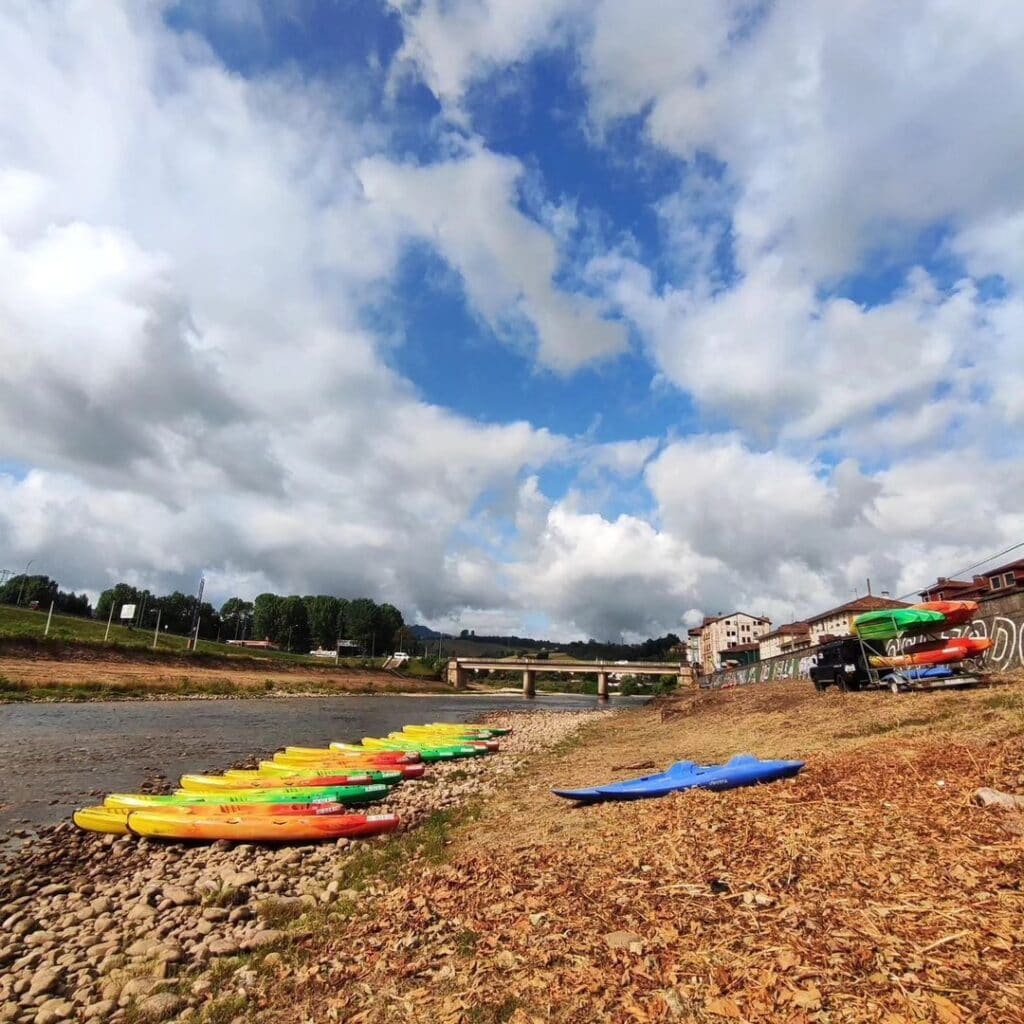 Fila de kayaks de colores alineados en la orilla de un río bajo un cielo parcialmente nublado, con un puente y edificios al fondo.