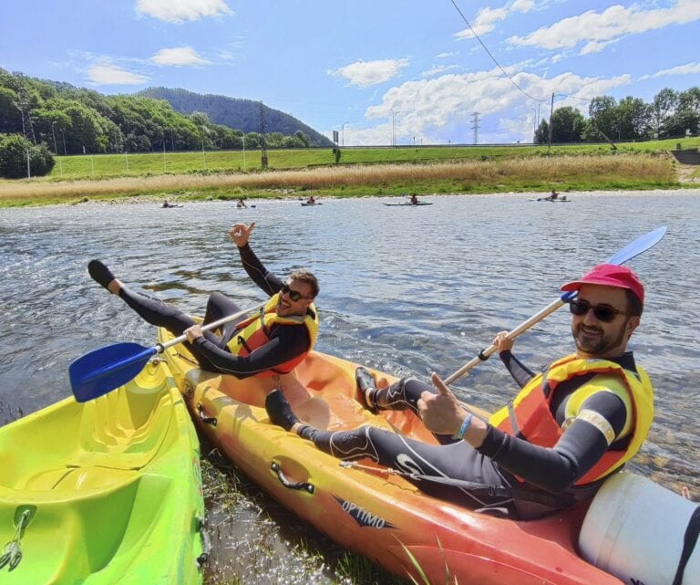 Dos personas con chalecos salvavidas y remos sentadas en un kayak junto a la orilla de un río, sonriendo y haciendo gestos de saludo.