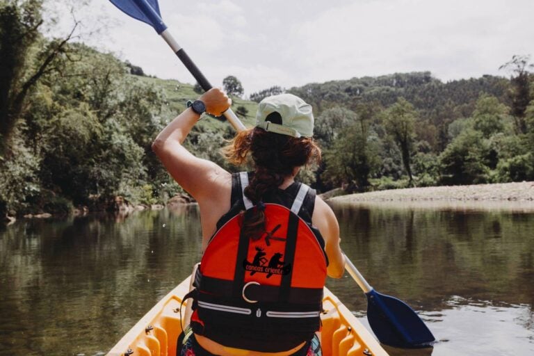 Persona remando en kayak con chaleco rojo en un río rodeado de árboles.