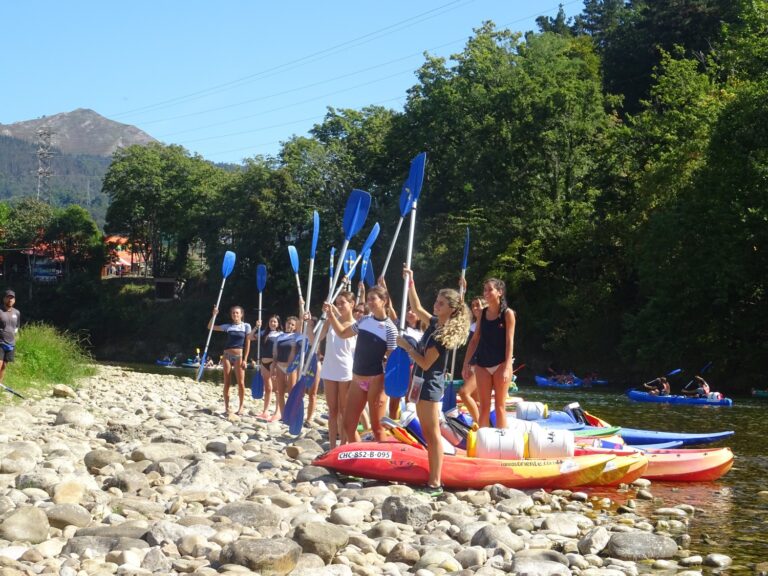 Grupo de jóvenes con remos azules en alto, junto a kayaks en la orilla rocosa de un río.